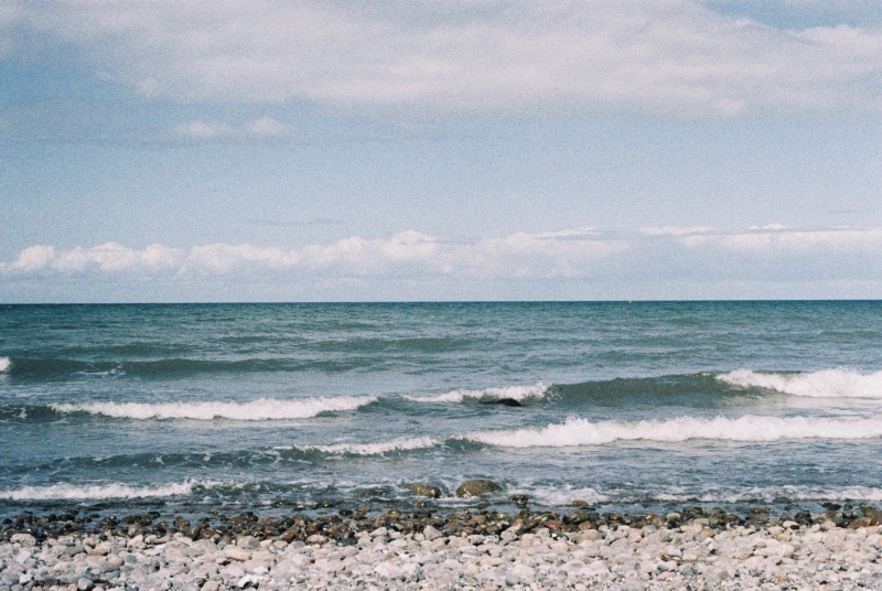Foto vom Strand aus aufgenommen, im Vordergrund Steine, dahinter das Meer, mit leichten, sich brechenden Wellen, im Hintergrund der Horizont mit wenigen Wolken