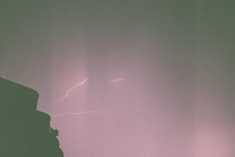 Aus dem Schatten eines Hauses am linken unteren Bildrand entspringen drei Blitze, die in den violetten Nachthimmel schießen.